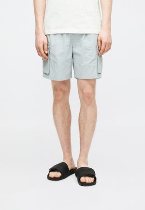 Persona che indossa pantaloncini cargo grigio chiaro e sandali neri a ciabatta, in piedi contro uno sfondo bianco semplice.