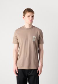 Ung mann iført en taupe kortermet T-skjorte med et lite abstrakt design og "BOSS"-tekst på brystet, kombinert med svarte bukser.