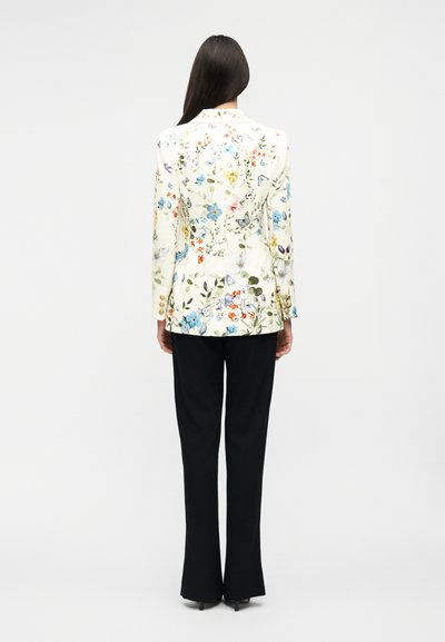 Γυναίκα με μακριά σκούρα μαλλιά, που φοράει ένα λευκό σακάκι με φυτικά μοτίβα και μαύρο παντελόνι, στέκεται με την πλάτη σε απλό φόντο.