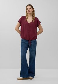 Bordeaux kortärmad blouse met een v-hals en een verstelbare trekkoord in de taille, gecombineerd met flared blauwe denim jeans en lichtgekleurde schoenen.