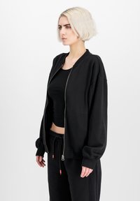Crna bomber jakna na zip od mekog materijala, s rebrastim manžetama i opuštenim krojem. Uparena s crnim skraćenim topom i trenirkama.