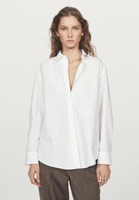 Camicia bianca con colletto e maniche lunghe, caratterizzata da una vestibilità comoda e un tessuto in cotone liscio. Abbinata a pantaloni a quadri marroni.