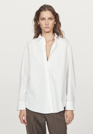 White button-up shirt with a collar and long sleeves, featuring a relaxed fit and smooth cotton fabric. Paired with brown checked trousers.