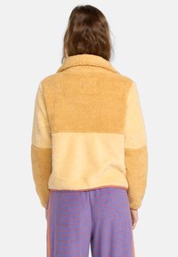 Fuzzy, color-blocked jacket in yellow and beige tones with a collar. Soft texture, cropped design, and a square label at the back.