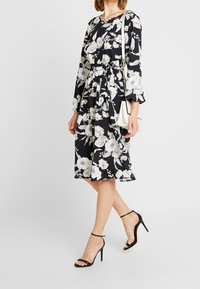 Floral dress in black fabric with white large flowers, long sleeves, tied waist, knee-length, paired with black strappy high heels.