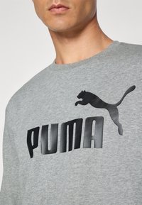 Homme portant un sweat-shirt gris avec un grand logo Puma noir et un graphique de puma bondissant sur la poitrine.