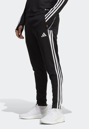 Osoba nosząca czarne spodnie dresowe Adidas z białymi trzema paskami i logo, w połączeniu z białymi butami sportowymi, stojąca na jednolitym tle.