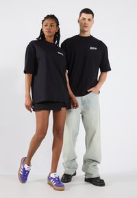  Sorte bomulds t-shirts med korte ærmer, der har et lille logo på brystet. Kvinden har en sort nederdel og lilla sneakers på; manden har lyse jeans på.