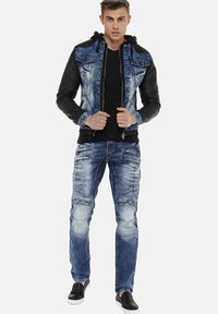 Denim jacket with black leather sleeves and hood, featuring multiple pockets. Worn over a black shirt and paired with distressed blue jeans and black shoes.