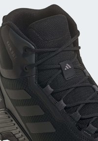 Gros plan d'une chaussure de sport noire avec tissu mesh, lacets texturés, col rembourré et trois bandes latérales. Texte "RAIN.RDY" visible au niveau de la cheville.