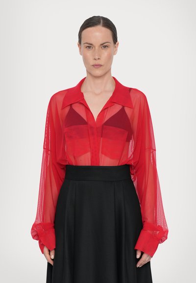 Blusa roja translúcida de manga larga con cuello, que presenta dos bolsillos en el pecho, combinada con una falda negra de talle alto. Textura suave.