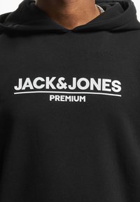 Sudadera negra con "JACK & JONES PREMIUM" en letras blancas en negrita en el pecho, llevada por una persona con una cadena de plata visible.