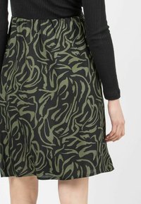 Ein schwarzes Kleid mit einem wirbelnden grünen Muster, das einen taillierten Oberteil und einen ausgestellt Rock hat. Der Stoff wirkt weich und leicht.