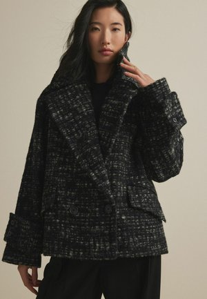 Manteau croisé noir et gris à motifs avec grands revers, couture visible et deux poches avant. Matériau en mélange de laine texturé.