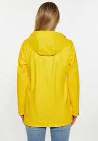 Vattentålig gul regnjacka med huva, slankt utformad, slät textur och kroppsnära silhuett. Har minimalistiska detaljer.