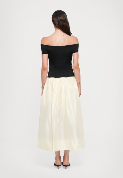 Sort off-shoulder top parret med en cremefarvet, knælang satin-nederdel med samlet talje og subtil glans, suppleret med sorte hæle.
