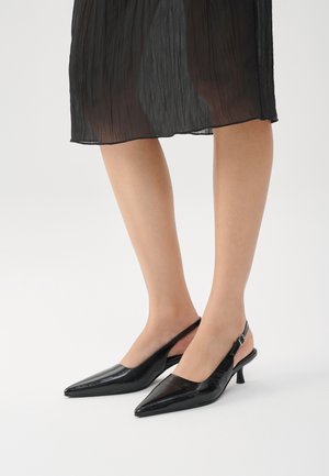 Jalat, joissa on musta läpikuultava midi-hame ja mustat kärjelliset slingback-kissakorkokengät, joissa on pieni solkinen hihna.