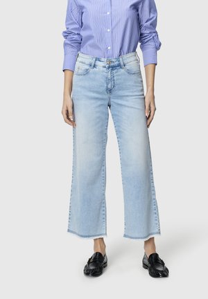 Kvinne iført lyseblå brede jeans, blå og hvit stripete skjorte med knapper, og svarte slip-on loafers, stående mot ensfarget bakgrunn.