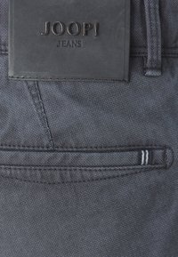 Ciemnoniebieskie dżinsy z denimu z czarną skórzaną naszywką z wytłoczonym logo marki "JOOP!", ozdobione subtelną teksturą w kratkę i białymi detalami szwów.