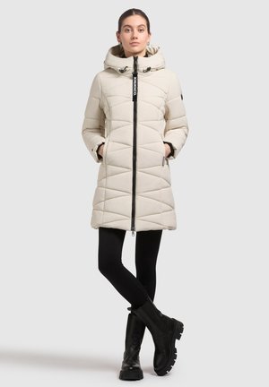 Beige quilted winter coat with a hood, featuring a black front zipper, and side pockets, worn with black leggings and chunky black boots.