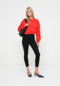 Sweatshirt rouge cropped à manches ballons, associé à un jean skinny noir taille haute avec cinq boutons, et des chaussures à talons aigus noires.