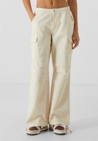 Beige cargo-bukser med løs pasform, der har to sidelommer, elastisk talje og blød bomullet tekstur. Komplementerer afslappet fodtøj.