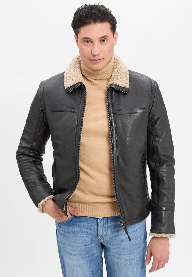 Schwarze Lederjacke mit einem beigen Lammfellkragen, Reißverschluss vorne und sichtbaren Steppnähten. Kombiniert mit einem beigen Rollkragenpullover und Jeans.