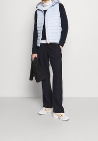 Personne portant un gilet matelassé bleu clair, un pull noir, un pantalon foncé, des baskets blanches avec des détails orange, tenant un sac matelassé noir.