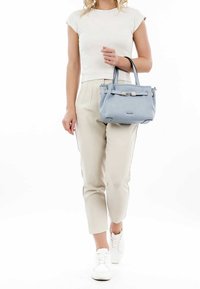 Žena v bílém triku a béžových kalhotách, držící světle modrou kabelku v pravé ruce, jde v bílých teniskách na bílém pozadí.