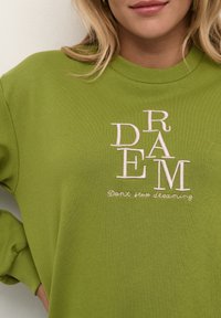 Sudadera verde de algodón con texto bordado en blanco "DREAM" con orientaciones mixtas de letras y la frase "No dejes de soñar" debajo.