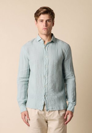 Giovane uomo con capelli castano chiaro indossa una camicia celeste chiaro a maniche lunghe con bottoni e pantaloni beige, in piedi davanti a uno sfondo beige uniforme.