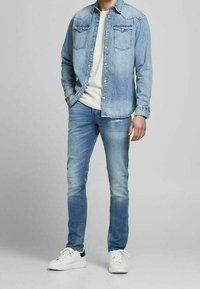 Camisa de mezclilla azul claro con dos bolsillos en el pecho, camiseta blanca debajo, jeans azules desgastados y zapatillas blancas con acentos negros.