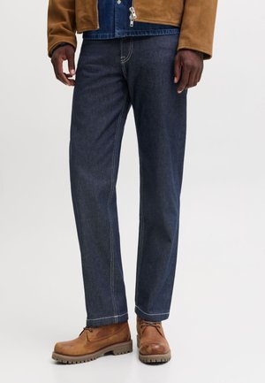 Homme portant un jean droit bleu foncé avec des coutures contrastantes et des bottes en cuir marron à lacets.