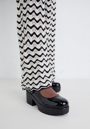 Tacones de plataforma de charol negro combinados con pantalones de punto con un patrón de zigzag en blanco y negro.