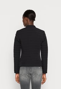 Zwarte gewatteerde jas met lange mouwen, staande kraag en textuurpanelen. G draagt met lichtgrijze jeans die een aansluitend ontwerp toont.