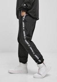Schwarze Sporthosen mit glänzender Textur, versehen mit einem weißen "STARTER"-Logo-Streifen an den Seiten, kombiniert mit weißen High-Top-Sneakern.