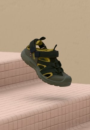 Une sandale d’extérieur noire et jaune, avec des brides réglables et une épaisse semelle grise, flotte au-dessus de marches carrelées de rose.