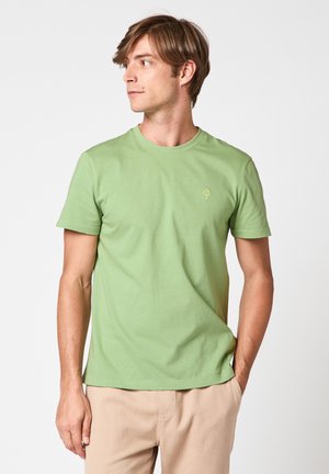 Jeune homme aux cheveux bruns courts portant un t-shirt à manches courtes vert clair et un pantalon beige, regardant vers sa gauche.