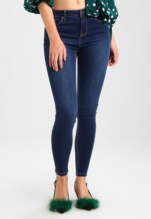 Femme portant un jean skinny bleu foncé, des talons pointus noirs avec des accents de fourrure verte, et un haut vert à pois noué à la taille.
