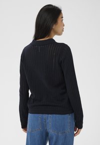 Hosszú, sötét hajú nő fekete bordás hosszú ujjú pulóverben és kék farmernadrágban háttal áll egy egyszínű világos háttér előtt.