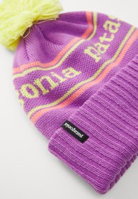 Gorro de malha roxo com um pom-pom amarelo. Apresenta um padrão riscas em rosa e amarelo, e uma manga canelada com uma etiqueta preta da Patagonia.
