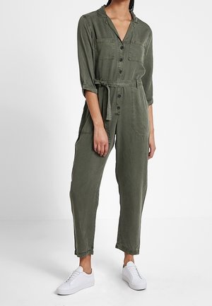 Kobieta w oliwkowozielonym kombinezonie roboczym z guzikami, z podwiniętymi rękawami, z paskiem w talii i białymi trampkami.