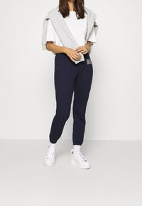 Marina sweatpants med ribbad mudd, med en logotyp på låret. I kombination med en vit t-shirt och en grå sweatshirt som är slängd över axlarna.