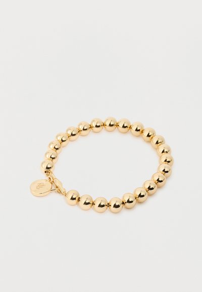 Bracelet en perles dorées avec des perles rondes polies et un petit charme circulaire près du fermoir sur un fond blanc.
