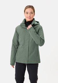 Giacca verde impermeabile con cappuccio, caratterizzata da una chiusura con zip e maniche regolabili. Indossata sopra una base nera, di consistenza liscia.