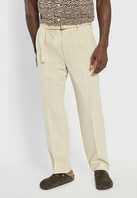 Pantalons plissés beiges avec une jambe droite, une ceinture assortie et des poches latérales, associés à des sabots en cuir marron foncé.