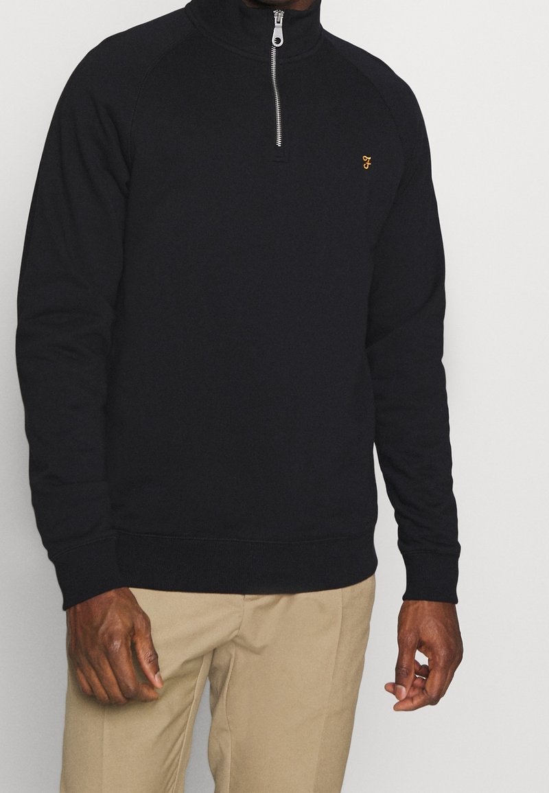 Sudadera negra con cremallera y cuello alto, con mangas raglán y un pequeño logo dorado. Combinada con pantalones beige.