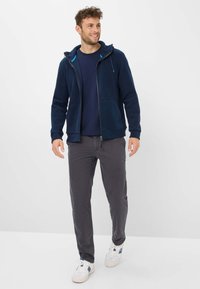 Felpa con cappuccio in pile blu navy con cerniera e due tasche, abbinata a una maglietta blu navy e pantaloni grigi. Le calzature hanno una base bianca con dettagli blu.