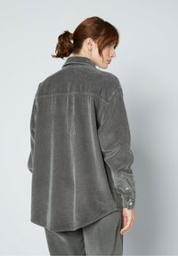 Szara koszula z kordurowego materiału o luźnym kroju, z kołnierzykiem, guzikami na mankietach i pionowym prążkowanym wykończeniu. Widok z tyłu ukazuje lekko zaokrąglony dół.