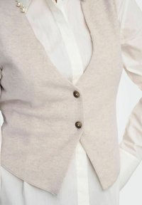 Gilet beige clair en tissu doux, avec un décolleté en V, deux boutons foncés et un ourlet arrondi, superposé à une chemise blanche.